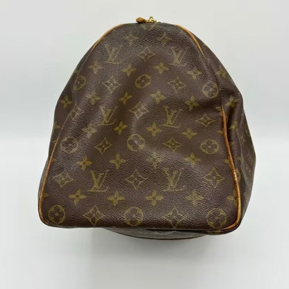 A side profile view of a Louis Vuitton monogram canvas travel duffle bag, highlighting the iconic interlocking LV pattern, structured dome shape, and honey-patina leather piping against a white background.⁠