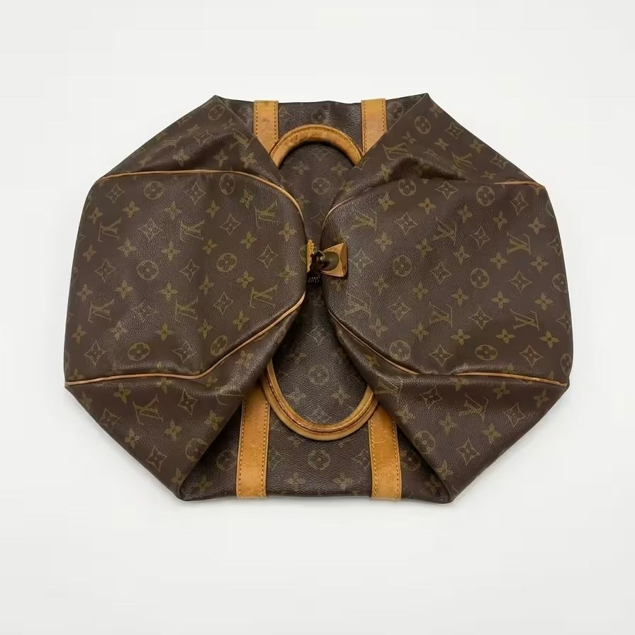 An overhead studio shot of a classic Louis Vuitton monogram canvas travel duffle bag folded completely flat, demonstrating its packable, space-saving design and authentic leather trim layout.⁠